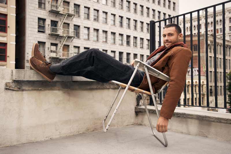 A masculine presenting person lounging on the rooftop of a building in an urban setting, wearing Koolaburra shoes.